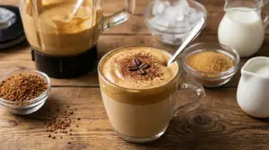 Crema al caffè servita in una tazza di vetro con cucchiaino, accanto a frullatore e ingredienti su tavolo di legno